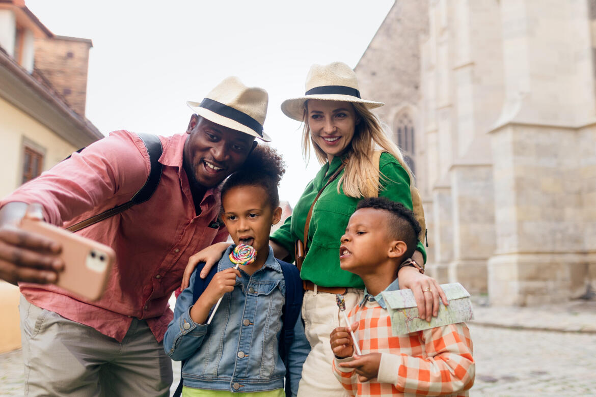 Happy family on vacation taking a selfie, featuring a smiling father, mother, and two children wearing hats and standing in front of historical buildings, illustrating the enjoyment of family stays offered by Appart'City.