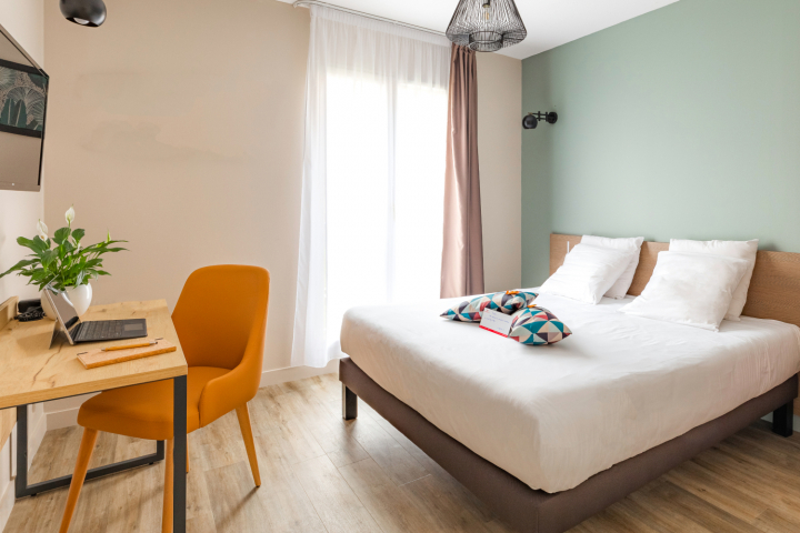 Comfort range hotel room with a double bed, white sheets, and patterned pillows, a wooden desk with an orange chair and a laptop, near a window with light curtains, creating a bright and soothing workspace.