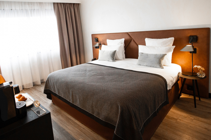 Elegantes Hotelzimmer der Collection-Reihe mit einem Kingsize-Bett, braunem Kopfteil, weißen Kissen, grauer Decke, Nachttisch mit einer Stimmungslampe, halb geöffneten Vorhängen, die Licht einlassen, und einem Höflichkeitstablett mit Gebäck.