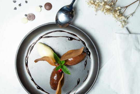 Vue de dessus d'un dessert artistique au chocolat disposé sur une assiette grise, avec une quenelle de glace vanille, une décoration en forme de feuille en chocolat, un coulis de chocolat dessinant une spirale et une petite feuille verte, accompagné d'une cuillère ancienne et de pétales séchés.