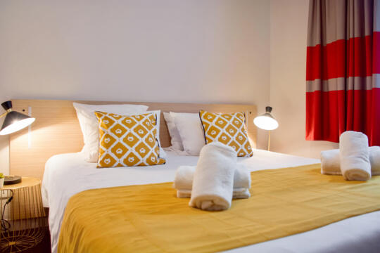 Welcoming hotel room with a king-size bed draped in a mustard yellow blanket and white pillows framed by two decorative cushions with yellow and white geometric patterns, two rolled white towels at the foot of the bed, wooden bedside tables with modern lamps, thick patterned red and grey curtains, a warm atmosphere and soft lighting