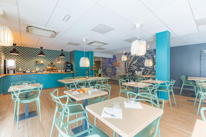 Salle de restaurant d'un appart'hôtel labellisé Clef Verte, avec des chaises et des tables en bois clair, des suspensions blanches modernes, un comptoir de bar carrelé, des murs bleu-gris et une fresque murale de feuillage, créant une ambiance contemporaine et écologique.