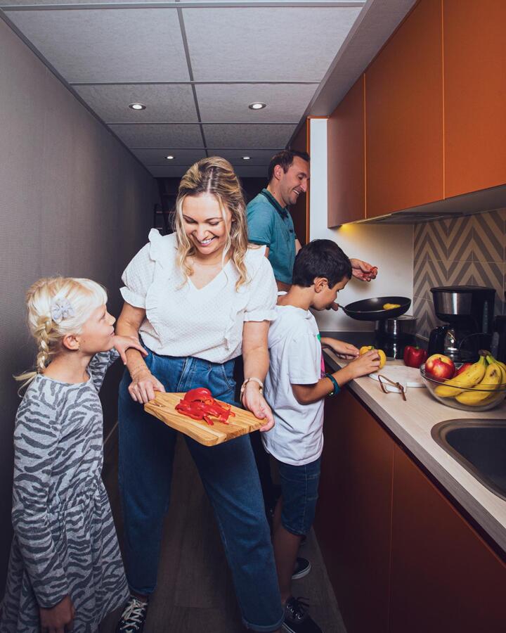 Happy family cooking together in an Appart'City apartment kitchen, with a mother showing peppers to her daughter, a son assisting in preparation, and a father cooking in the background, highlighting the comfort of family stays at Appart'City.
